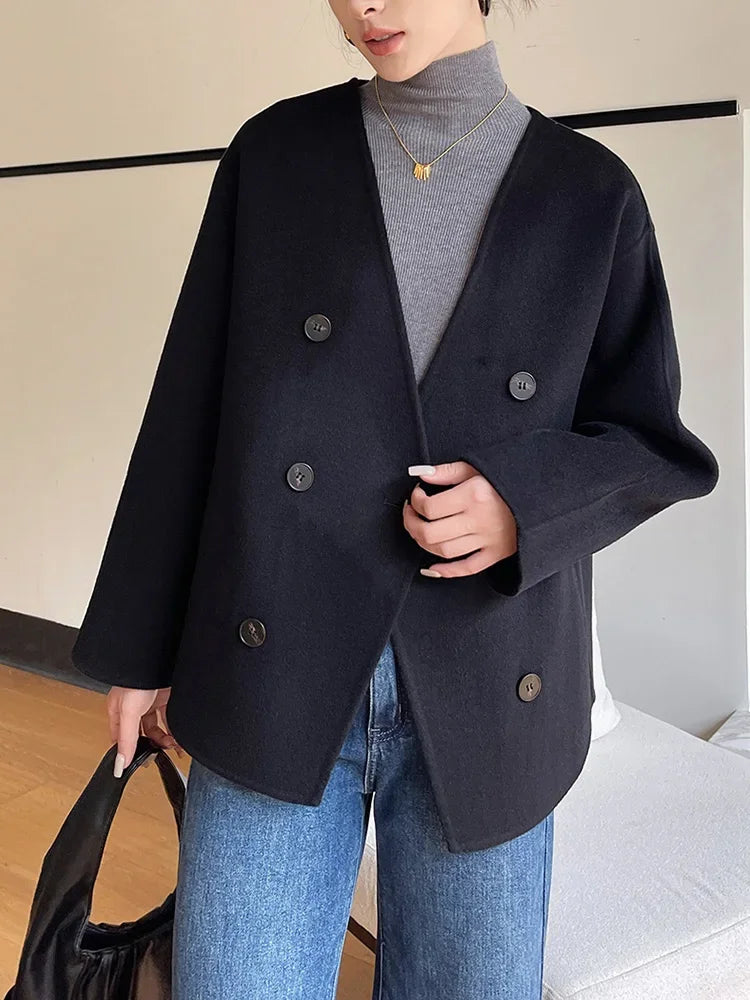 Person wearing a black double-breasted coat over a gray turtleneck and blue jeans, holding a black bag indoors.