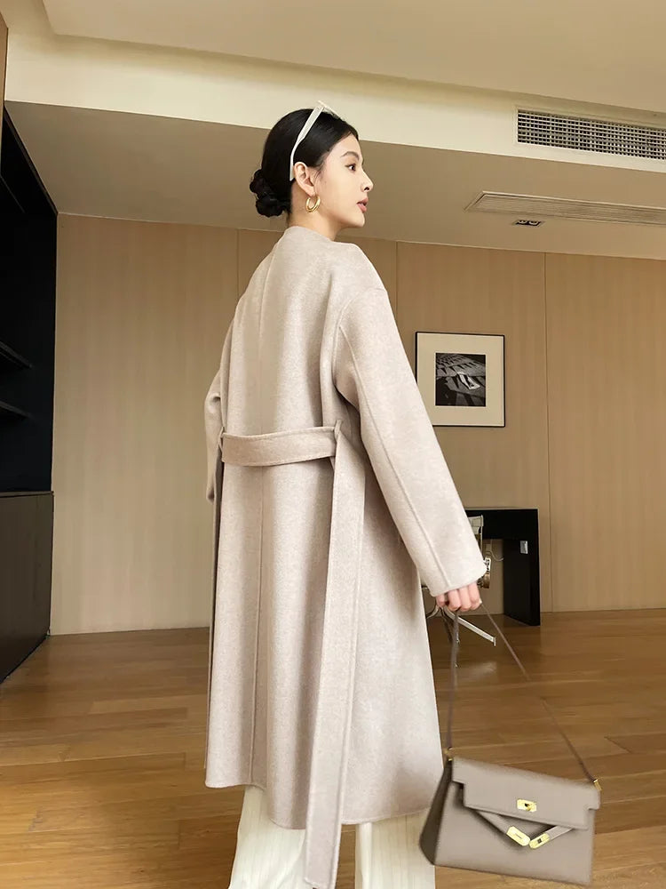 Woman in a long beige coat holding a handbag in an indoor setting.