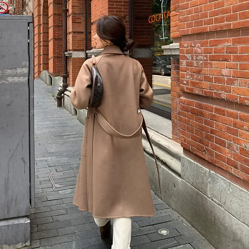 Woman in a beige coat walking on a sidewalk next to a brick building.