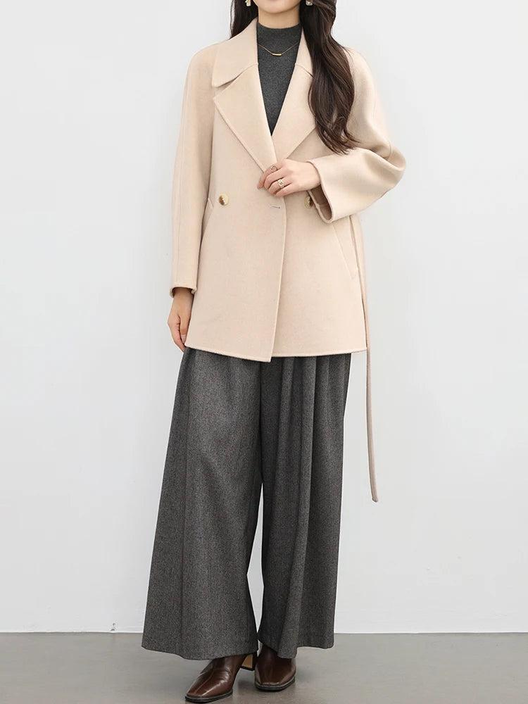 Person wearing a beige coat and gray wide-leg pants against a white background