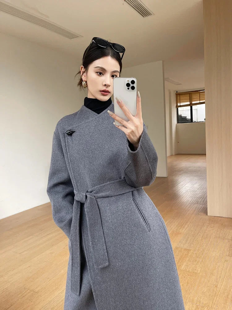 Woman taking a mirror selfie wearing a gray coat in a minimalistic room.