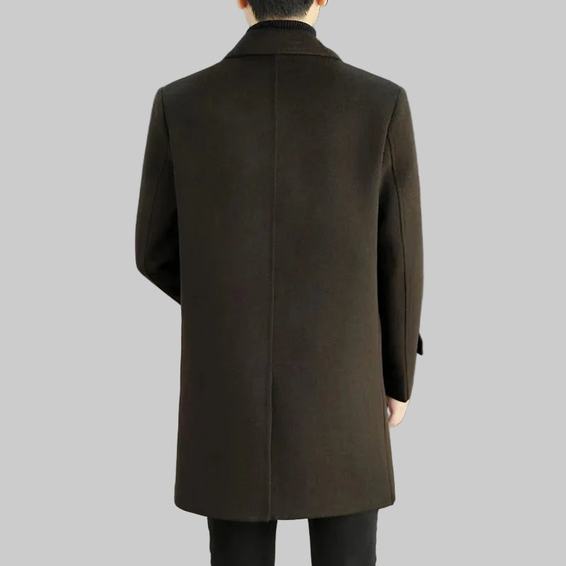 Person wearing a long dark coat in an indoor setting with a white sofa and gray wall.