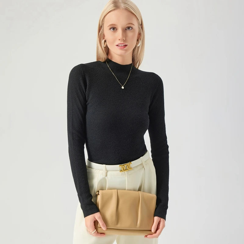 Woman holding a beige handbag wearing a black turtleneck and white pants on a light gray background