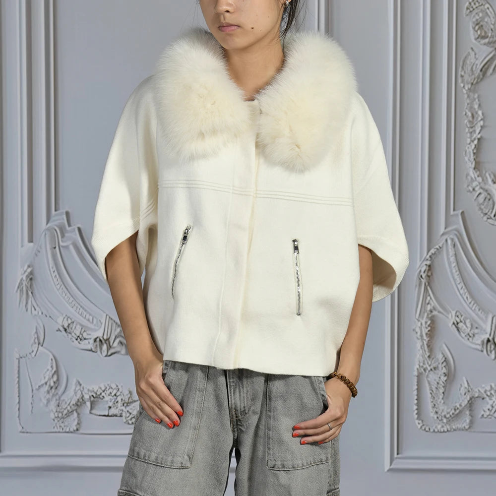Person wearing a white poncho with a fur collar against an ornate gray wall.