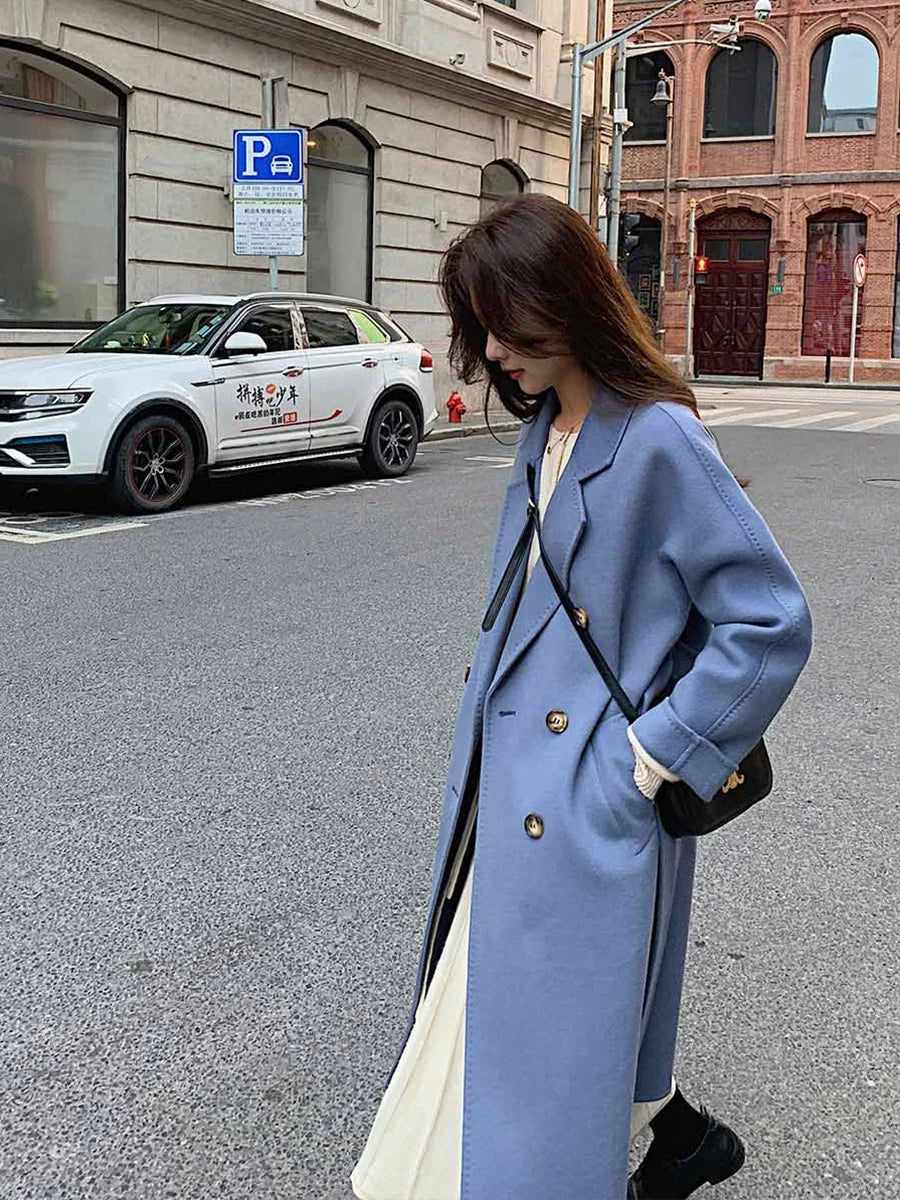 Woman in a blue coat walking on a city street with a car and building in the background.