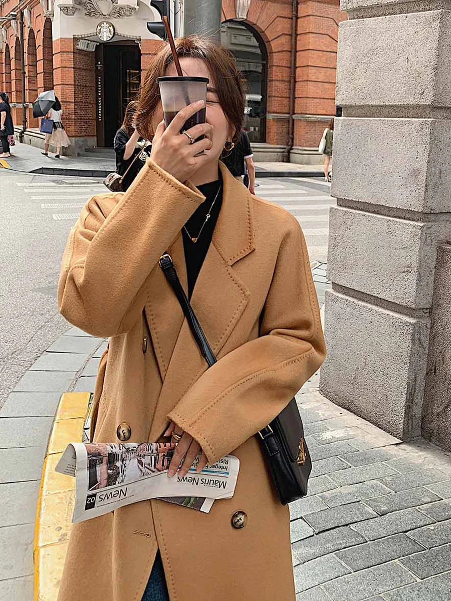 Person in a tan coat holding a phone and newspaper on a city street.