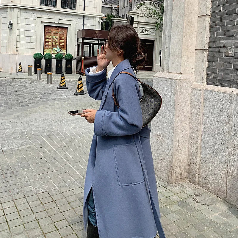Woman in a blue coat walking on a city street, holding a phone.